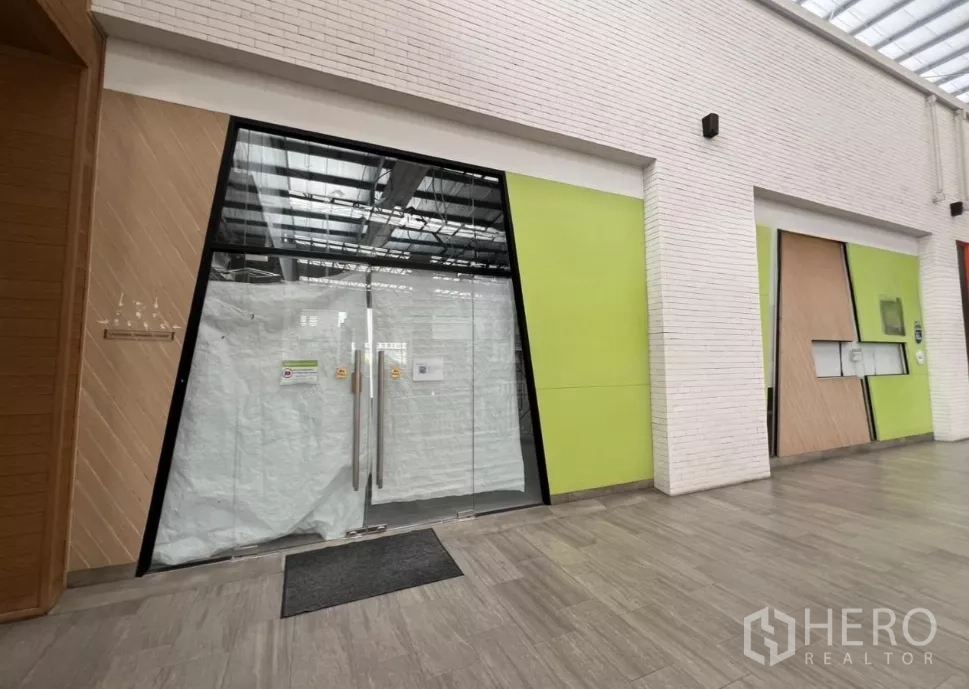 零售空间 出租在Khlong Toei, Bangkok - 社区商场走廊，现代店面，宽幅玻璃双开门与浅绿色面板。