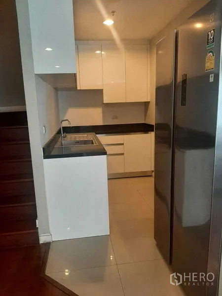 Condominium for rent or sale in Huai Khwang, Bangkok - Modern kitchenette with glossy white cabinets, sink, and stainless refrigerator.
