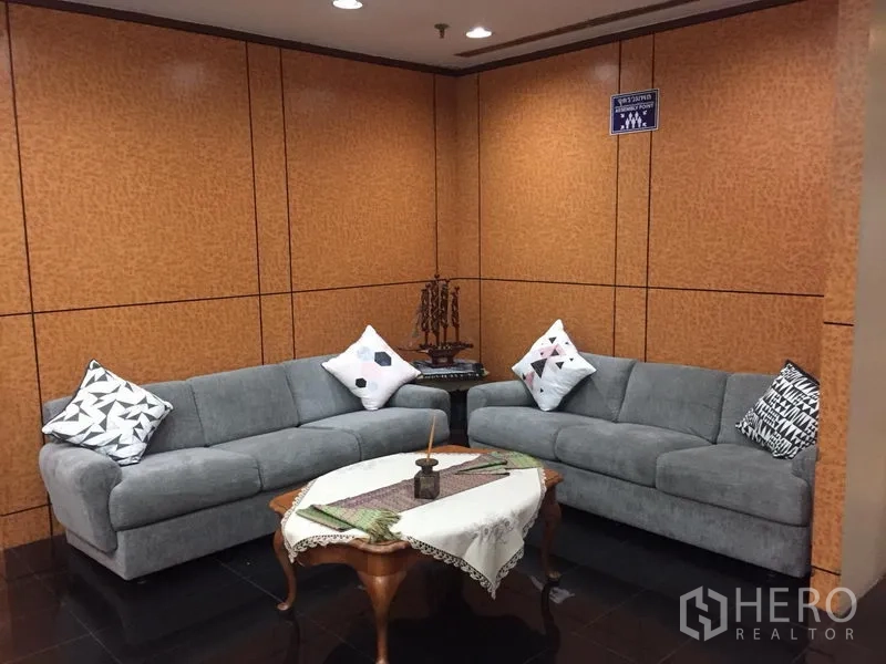 สำนักงาน ให้เช่าในRatchathewi, Bangkok - มุมรับรองแขกพร้อมโซฟาสีเทา 2 ตัว โต๊ะกาแฟไม้ปูผ้าลูกไม้ และผนังลายไม้ภายใต้ไฟดาวน์ไลต์