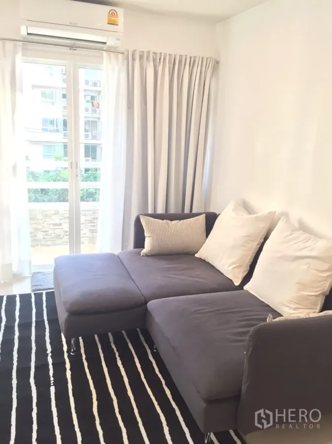 Condominium for rent in Watthana, Bangkok - Bright living room with gray sectional sofa, striped rug, air conditioner, and sliding doors opening to a small balcony.