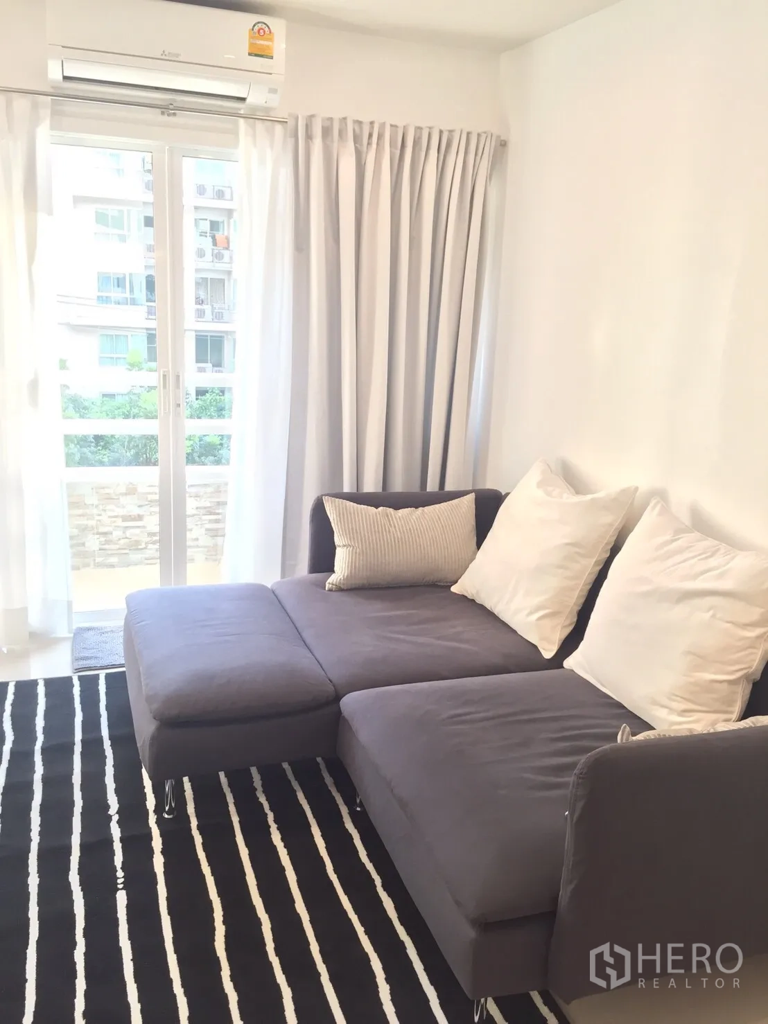Condominium for rent in Watthana, Bangkok - Bright living room with gray sectional sofa, striped rug, air conditioner, and sliding doors opening to a small balcony.