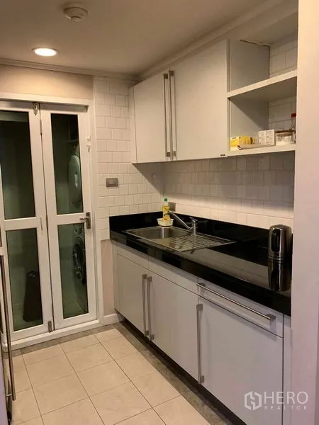 Condominium for rent in Pathum Wan, Bangkok - Galley kitchen with white cabinets, black countertops and door to a utility area.