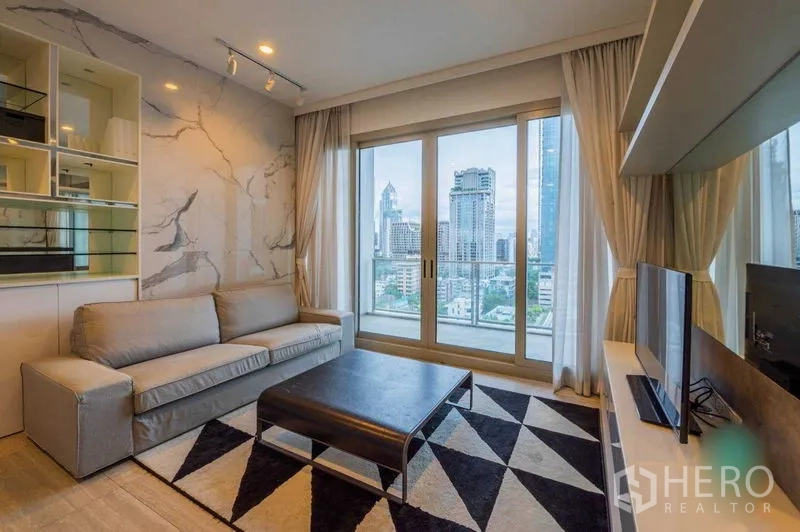 Condominium for rent in Pathum Wan, Bangkok - Living room with gray sofa, TV console, geometric rug, and floor-to-ceiling windows overlooking the Bangkok skyline.