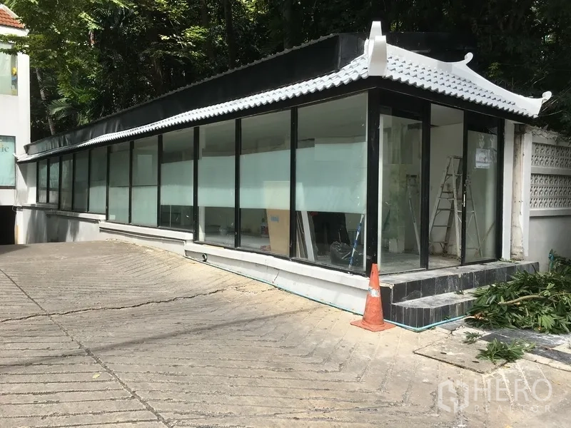 零售空间 出租在Watthana, Bangkok - 转角玻璃店面，黑白屋檐与连续橱窗，带小台阶与斜坡通道，绿植环绕。