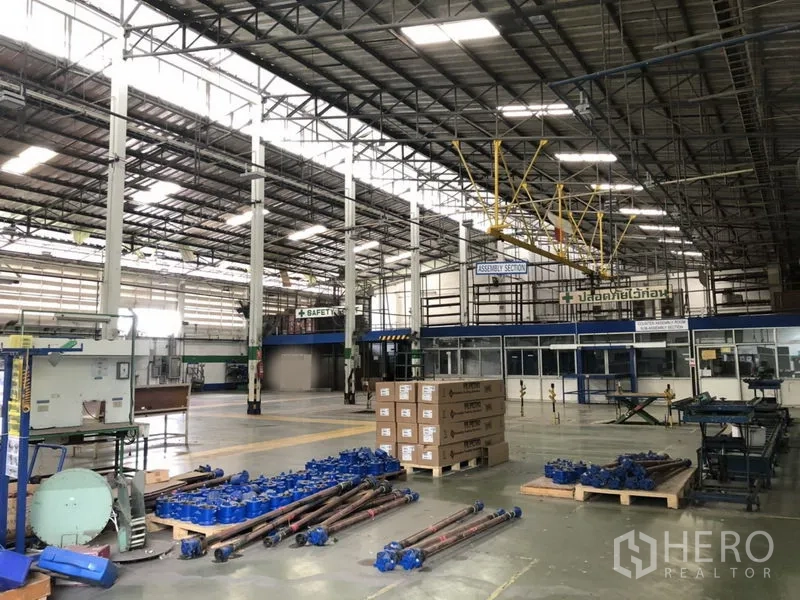 Factory for rent in Lam Luk Ka, Pathum Thani - Assembly section showing pallets, equipment and a glass‑fronted office under high clearance roof and cranes.