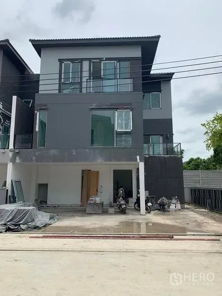 Detached House for sale in Prawet, Bangkok - Three-storey modern detached house with dark gray facade, large windows, balconies, and a covered parking area at the front.