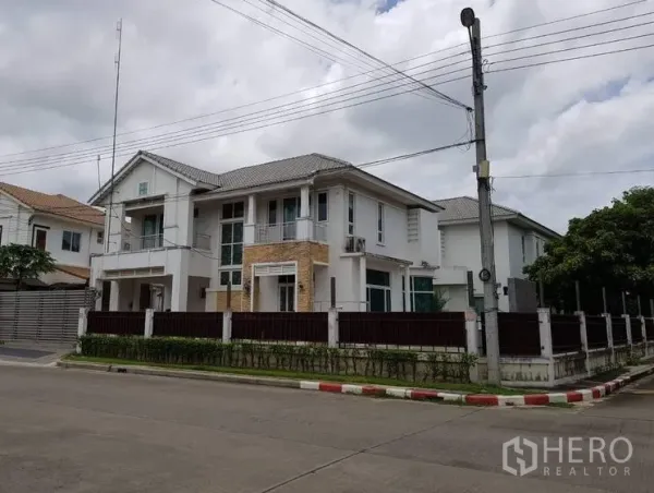 บ้าน ขายในLat Krabang, Bangkok - มุมมองแปลงมุมของบ้านเดี่ยวสไตล์โมเดิร์น สีขาว พร้อมรั้วรอบขอบชิด ไม้พุ่ม ระเบียง และหลังคากระเบื้อง บนถนนหมู่บ้านเงียบสงบ