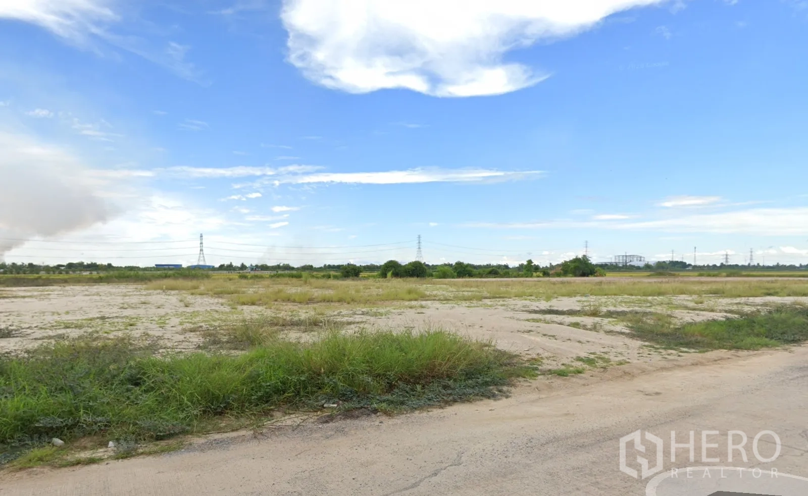 Land for sale in Sai Noi, Nonthaburi - Cleared open plot beside a paved road with grass patches and distant power lines.