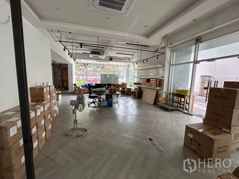 Office Space for rent in Khlong Toei, Bangkok - Ground‑floor open‑plan office with floor‑to‑ceiling windows, herringbone flooring, exposed track lighting and stacked boxes.