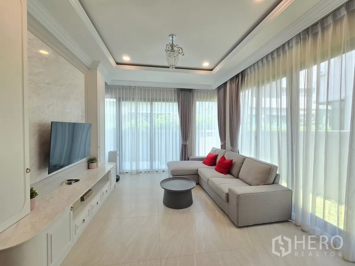 Detached House for rent in Prawet, Bangkok - Bright living room with corner sofa, floor‑to‑ceiling sheer curtains and a built‑in TV wall.