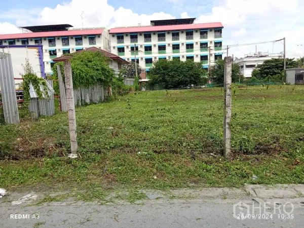 Land for rent in Khan Na Yao, Bangkok - Level green field inside a wire fence facing a local road and apartment buildings.