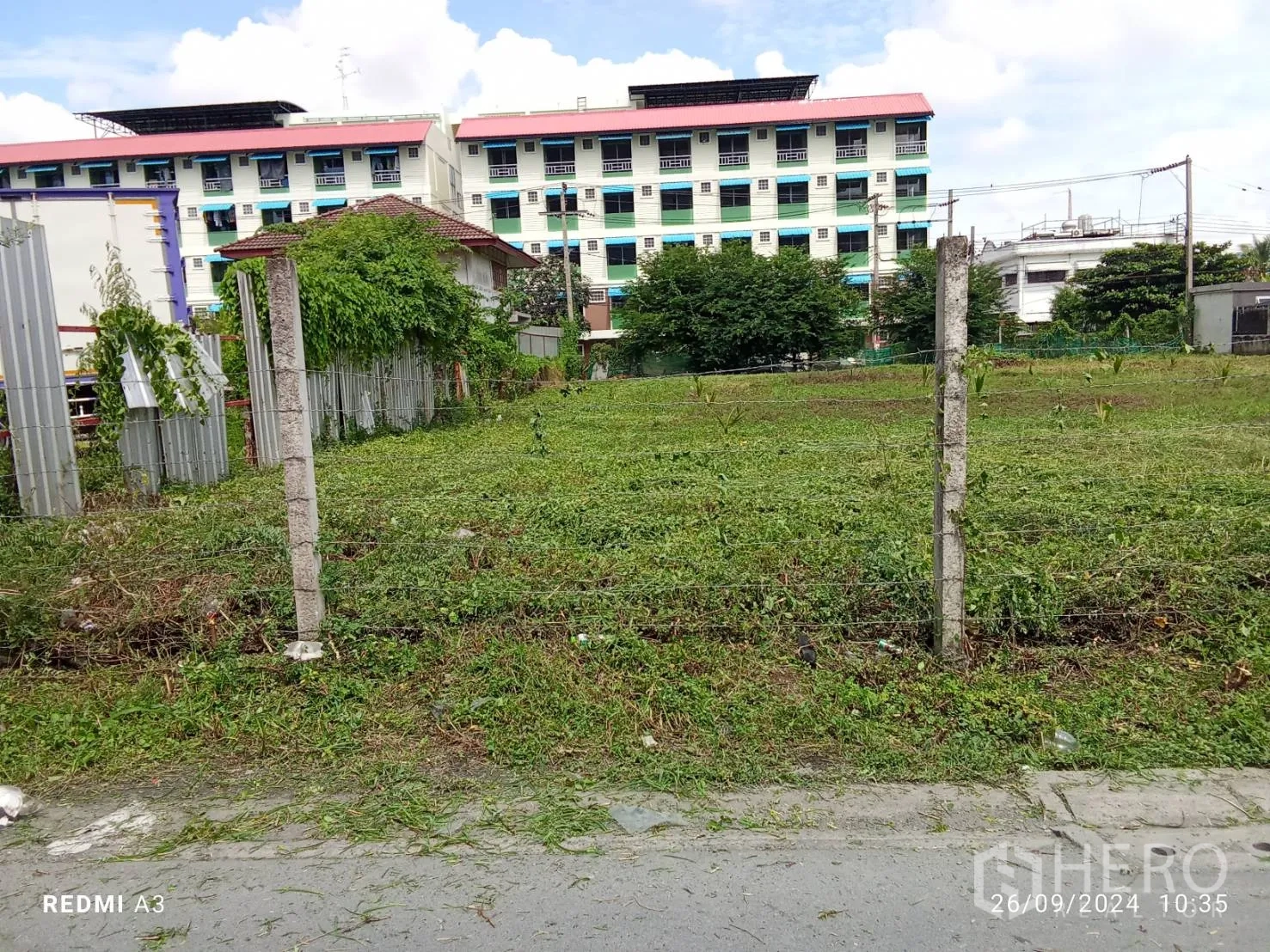 Land for rent in Khan Na Yao, Bangkok - Level green field inside a wire fence facing a local road and apartment buildings.