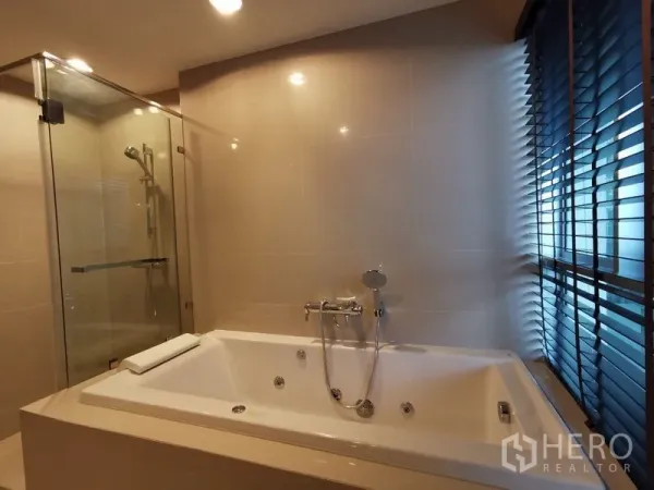 Condominium for rent or sale in Huai Khwang, Bangkok - Bathroom with a large jacuzzi bathtub, glass-enclosed shower and window blinds.