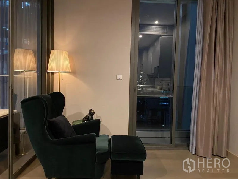 Condominium for rent in Huai Khwang, Bangkok - Cozy reading corner with armchair and ottoman beside a floor lamp and sliding balcony door at night.