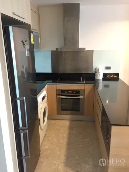Condominium for rent in Sathon, Bangkok - Modern kitchen with black countertops, built-in oven, cooktop, range hood, fridge and washing machine.