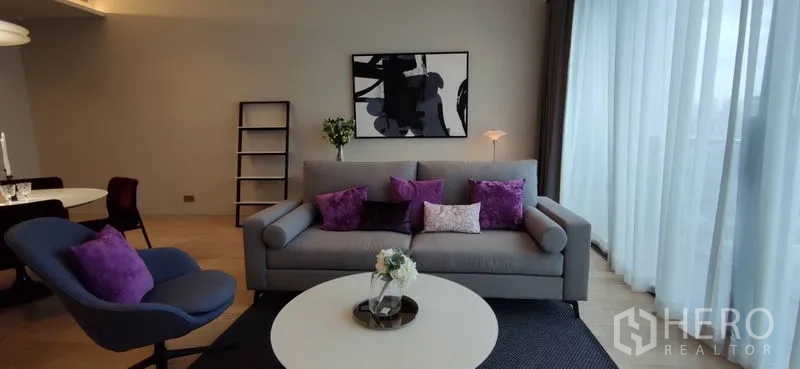 คอนโด ให้เช่าหรือขายในWatthana, Bangkok - ห้องนั่งเล่นโทนสบาย โซฟาสีเทาพร้อมหมอนสีม่วง และหน้าต่างบานใหญ่ผ้าม่านโปร่ง