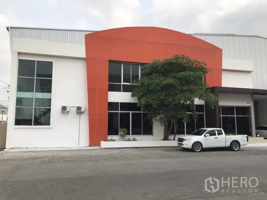 工厂 出租在Samut Prakan - 现代化工厂外立面，橙色门头与两层玻璃办公室及车道。
