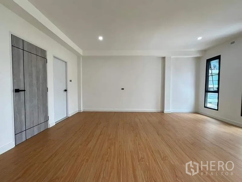 Townhouse for sale in Watthana, Bangkok - Open-plan room with wood floors, white walls, twin interior doors and a corner window.