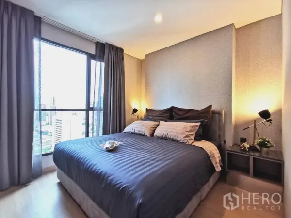 Condominium for rent in Huai Khwang, Bangkok - Master bedroom with large window, city skyline view, grey curtains and bedside lamps in a modern condominium.