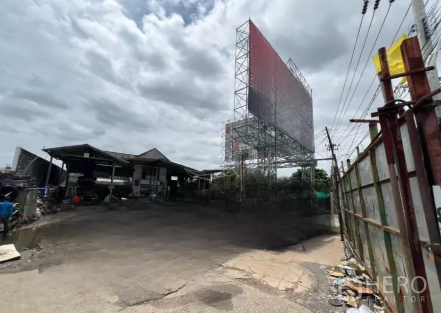 Land for sale in Bang Kapi, Bangkok - Paved yard with tall billboard structure, small shed, boundary fencing and power lines under a cloudy sky in Bang Kapi.