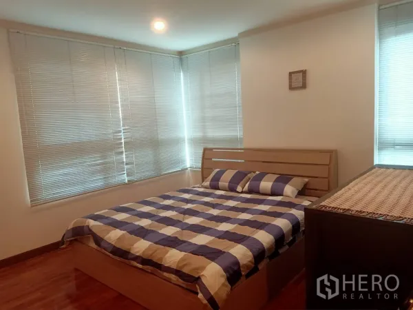 Condominium for rent or sale in Watthana, Bangkok - Corner bedroom with large windows and blinds, wooden bed with striped bedding and warm wood flooring.