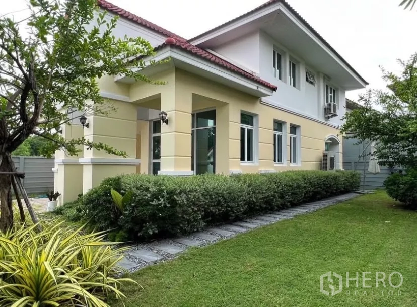 Detached House for rent or sale in Prawet, Bangkok - Corner view of a two-storey detached house with yellow facade, red tile roof and manicured lawn.