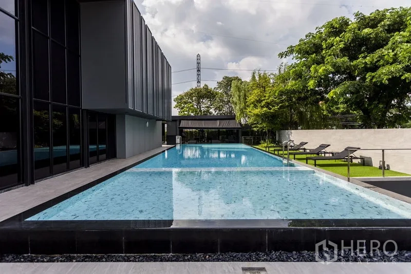 联排别墅 出租在Samut Prakan - 社区长条形泳道泳池，旁为现代会所与躺椅。