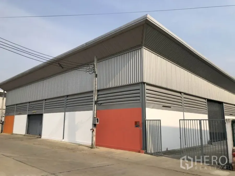 Warehouse for rent in Sai Noi, Nonthaburi - Corner view of a metal-clad warehouse with roller door and gated entrance on a concrete road.