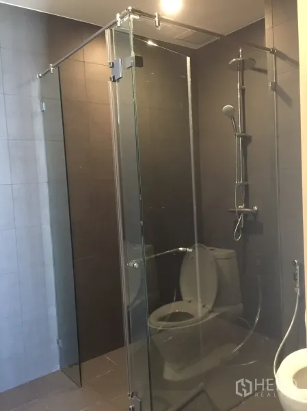 คอนโด ขายในPathum Wan, Bangkok - ห้องน้ำสมัยใหม่พร้อมตู้กระจกกั้นอาบน้ำและฝักบัวเรนชาวเวอร์บนผนังโทนเข้ม.