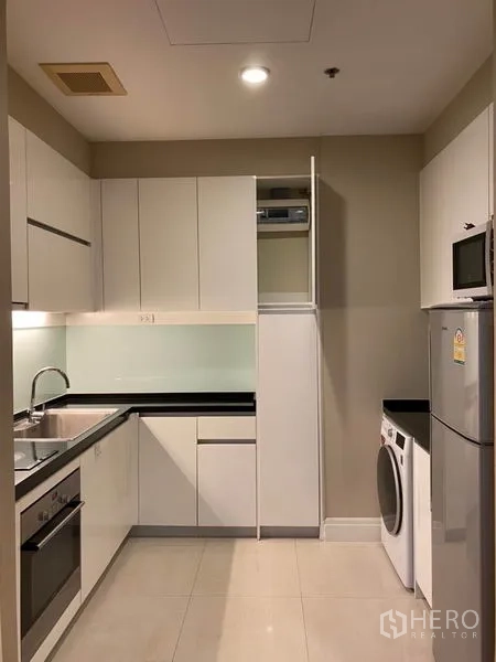 Condominium for rent in Khlong Toei, Bangkok - Modern L-shaped kitchen with white cabinets, built-in oven, fridge and washing machine.