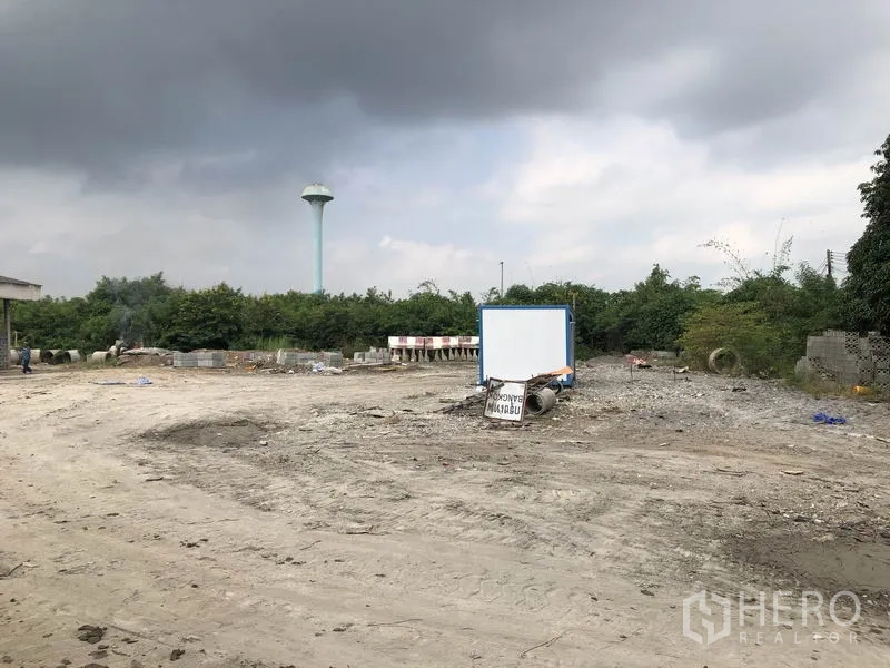 工厂 出租在Phra Nakhon Si Ayutthaya - 开放工业用地，地面为碎石，设有告示牌，背景有水塔与绿化。