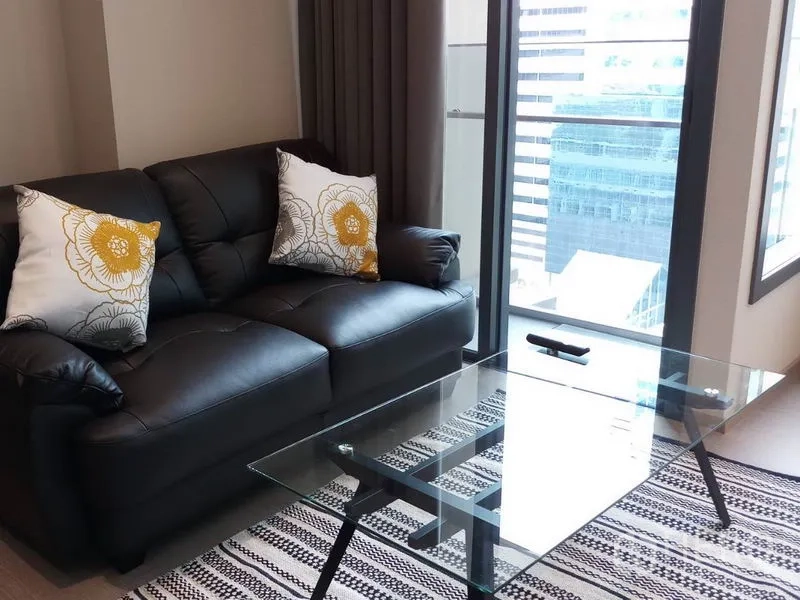 公寓 出租在Khlong Toei, Bangkok - 采光良好的客厅，黑色皮沙发、花纹抱枕与玻璃茶几，窗外城市景观