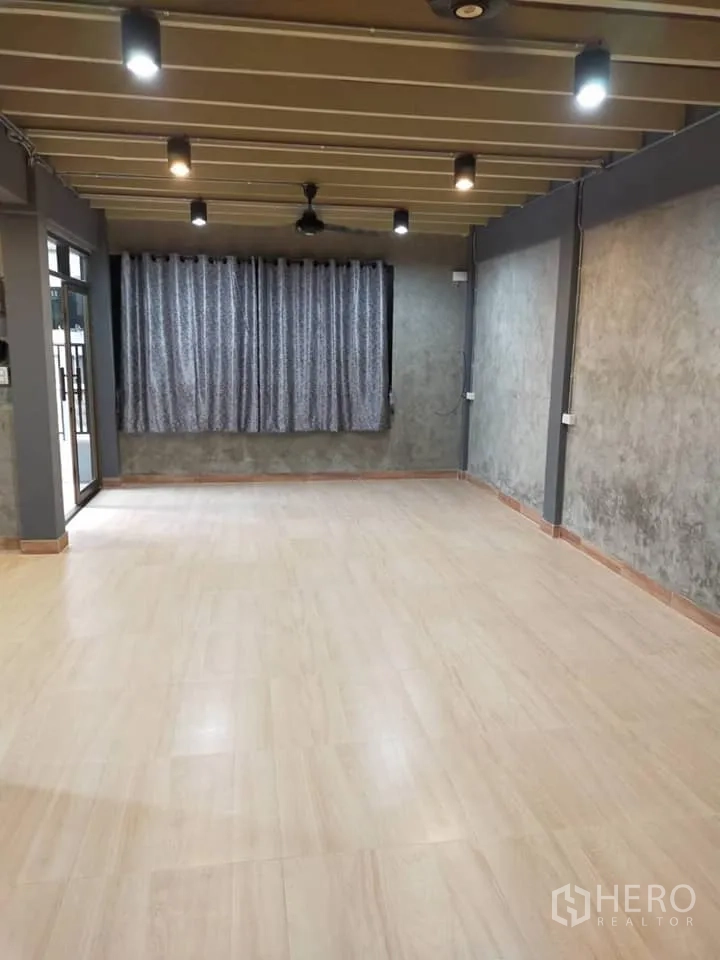 Detached House for rent in Watthana, Bangkok - Spacious living room with light wood floors, exposed beam ceiling and LED spotlights, featuring a large curtained window.