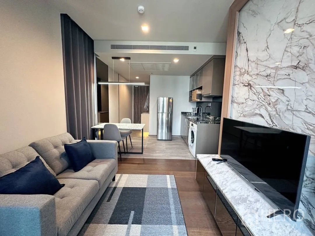 Condominium for rent in Khlong Toei, Bangkok - Modern living-dining area with gray sofa, marble TV wall, flat-screen TV, open kitchen with fridge and washing machine, and a dining table with pendant light.