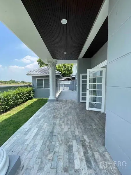 Detached House for sale in Bang Na, Bangkok - Covered front terrace with stone tiles, white columns and French doors beside a neat lawn.
