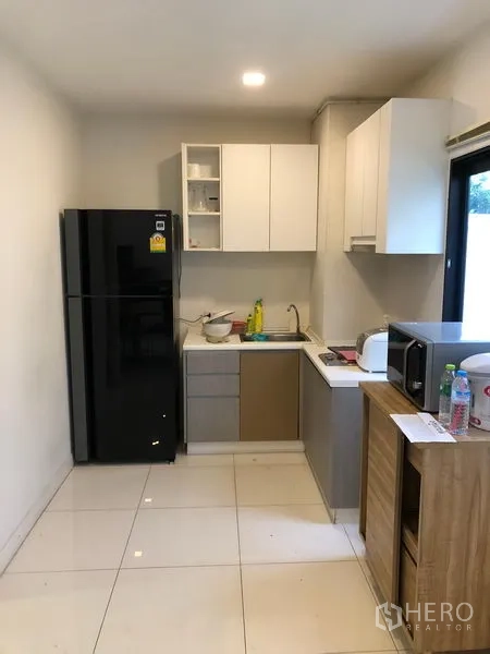 Townhouse for rent or sale in Saphan Sung, Bangkok - Compact kitchen with white cabinetry, black refrigerator, sink and side window.