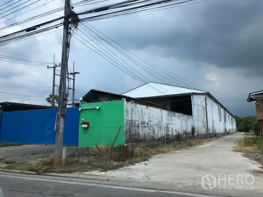 คลังสินค้า ให้เช่าในSam Khok, Pathum Thani - ภายนอกโกดังหลังคาสูง พร้อมประตูทางเข้า รั้วรอบขอบชิด และทางคอนกรีตกว้าง ในอำเภอสามโคก ปทุมธานี ใต้ท้องฟ้าครึ้ม
