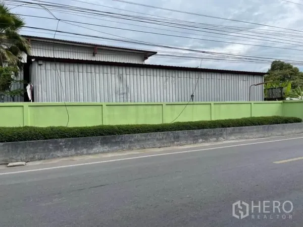 货栈 出售在Lam Luk Ka, Pathum Thani - 轻绿色围墙后的长条金属板仓库，沿安静的双车道公路。