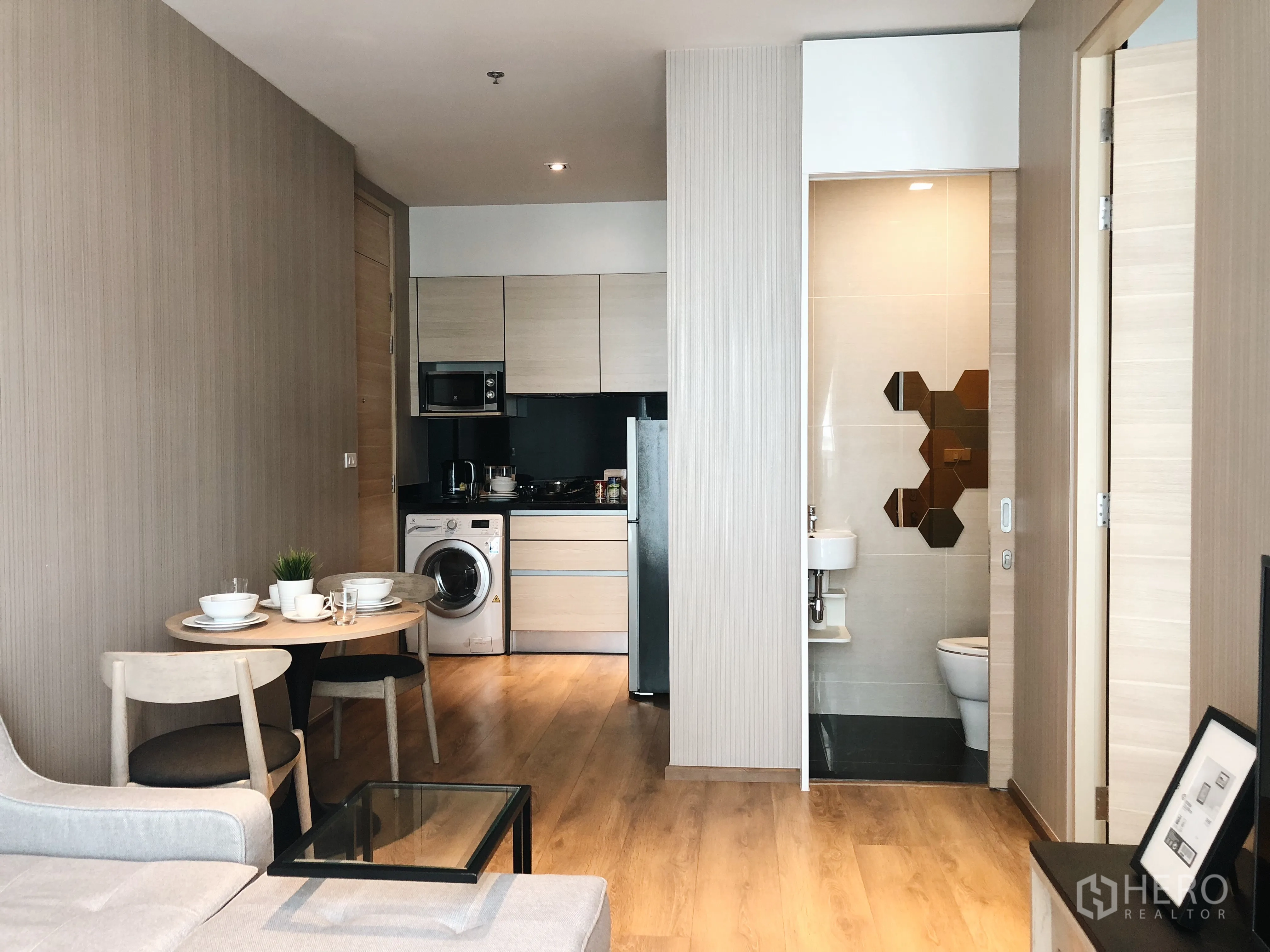 Condominium for rent in Khlong Toei, Bangkok - Open-plan living with fitted kitchen, dining set for two, microwave, fridge, washing machine, and bathroom entrance.