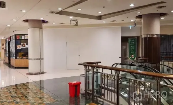 零售空间 出租 - 购物中心走廊，空置白色店面，旁有扶梯栏杆与光亮地面。
