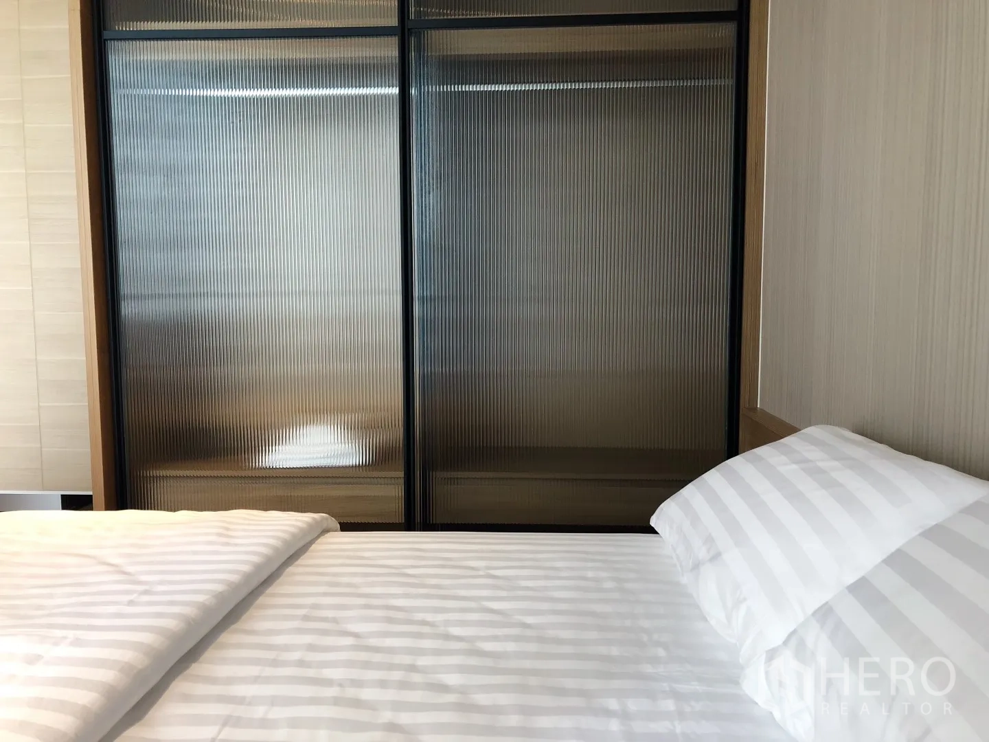 Condominium for rent in Khlong Toei, Bangkok - Cozy bedroom with striped bedding and ribbed glass sliding wardrobe doors.
