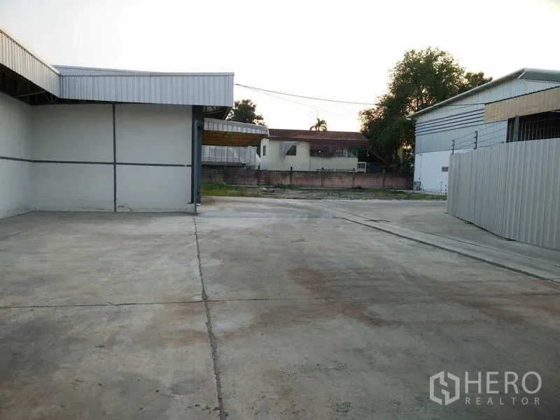 Warehouse for rent in Suan Luang, Bangkok - Spacious concrete yard between warehouse buildings with perimeter fence and houses beyond.
