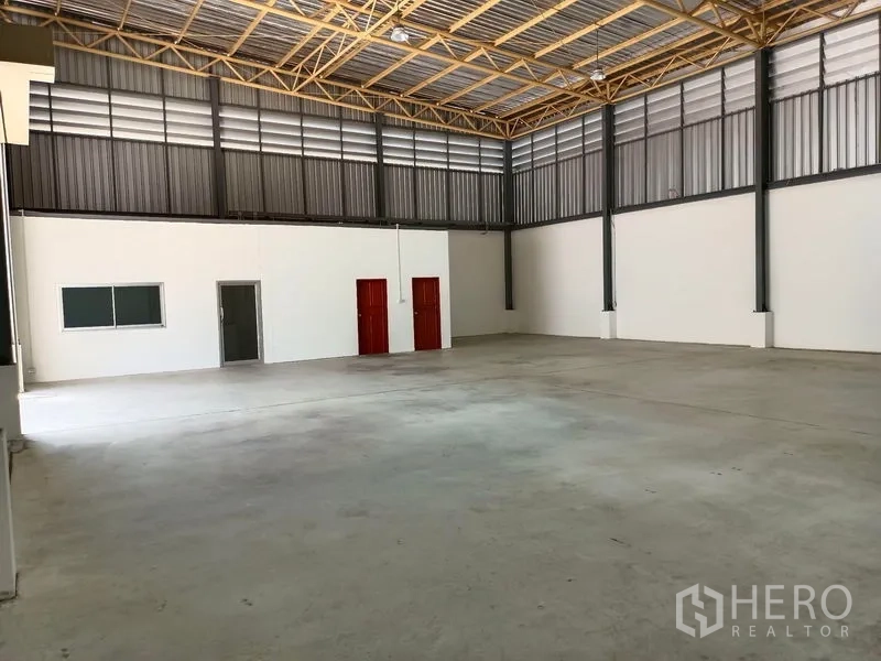 Warehouse for rent in Suan Luang, Bangkok - Large warehouse bay with truss roof, white partitioned office, and two red service doors.