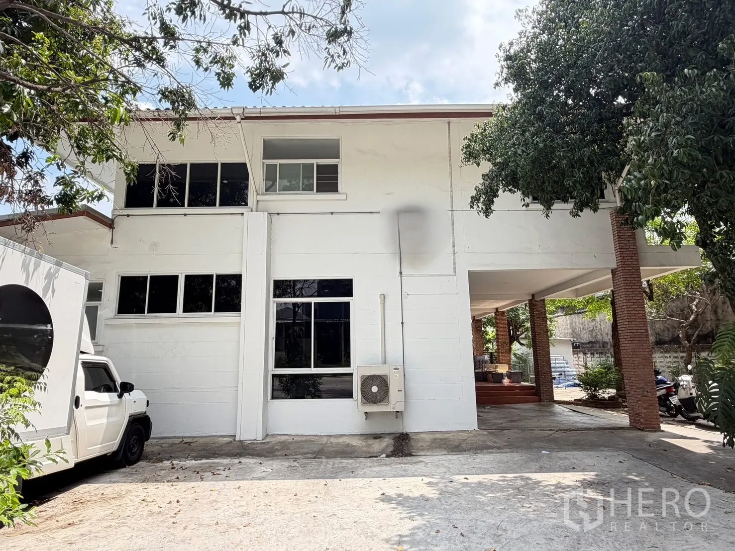 Detached House for rent in Watthana, Bangkok - Front exterior of a two-storey white detached house with brick columns, driveway, and mature trees.