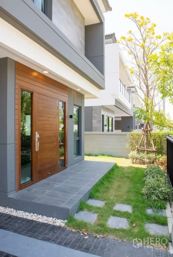 Detached House for rent in Bang Phli, Samut Prakan - Front entrance with wooden door, tiled porch and a small lawn with shrubs.
