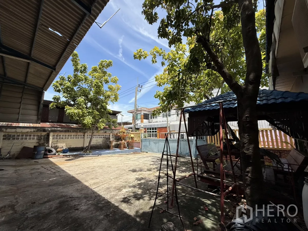 Warehouse for sale in Phra Samut Chedi, Samut Prakan - Spacious concrete yard with trees, a small pavilion and front sliding gate under clear sky.