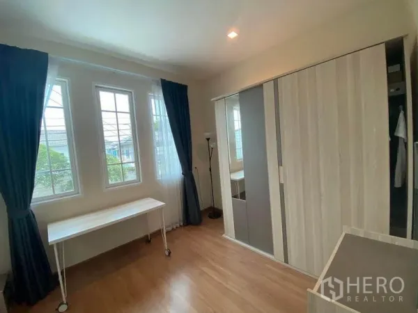 Townhouse for sale in Saphan Sung, Bangkok - Sunlit bedroom with large windows, dark curtains, a white rolling desk and a sliding wardrobe on wood flooring.