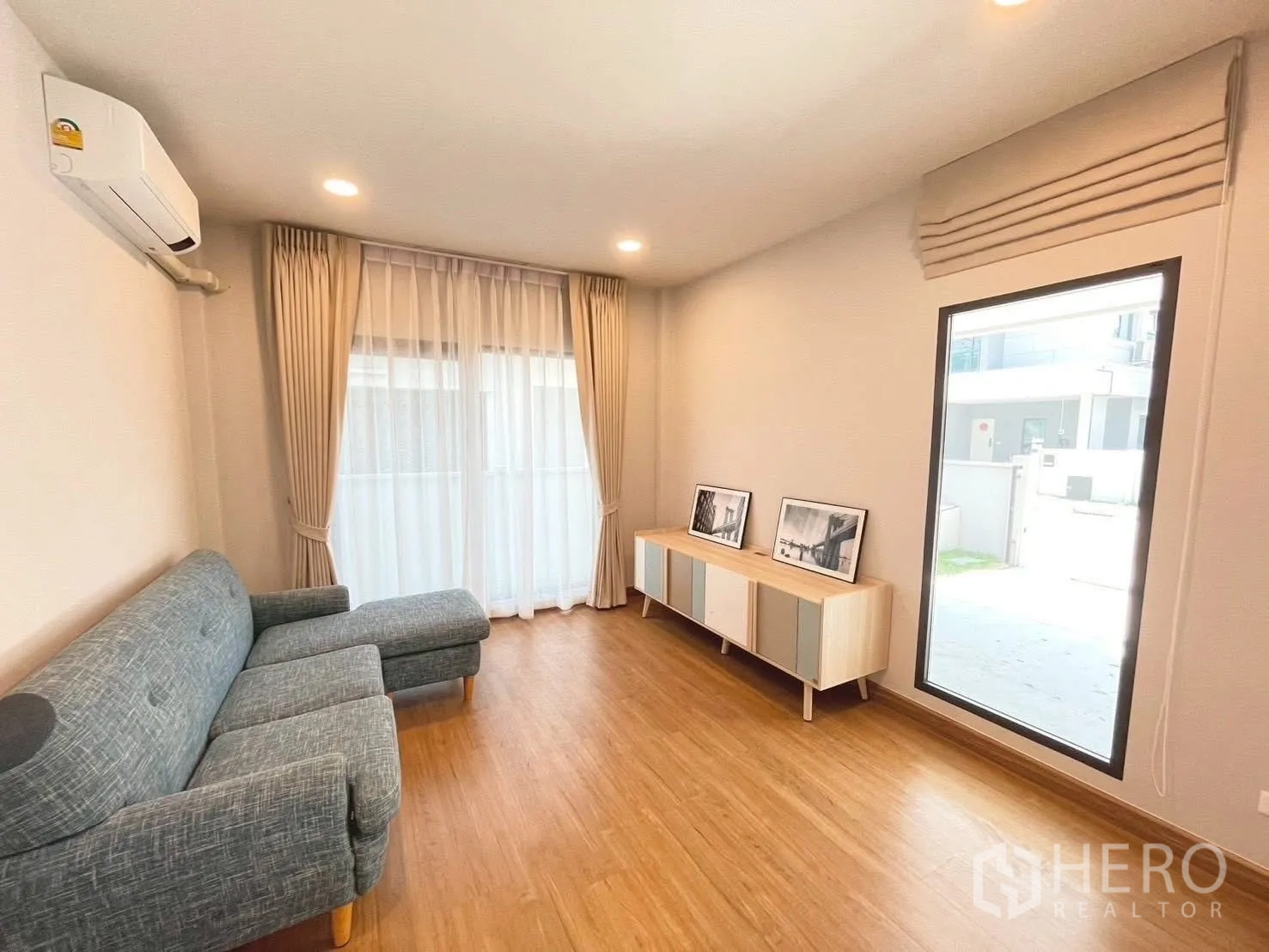 Detached House for rent in Bang Phli, Samut Prakan - Bright living room with gray sofa, wood floor, and TV console by large windows.