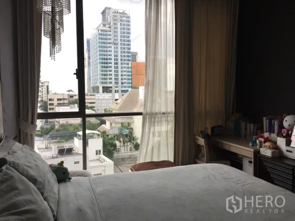 Condominium for sale in Watthana, Bangkok - Cozy bedroom with bed, study desk, sheer curtains, and floor-to-ceiling window overlooking the city.