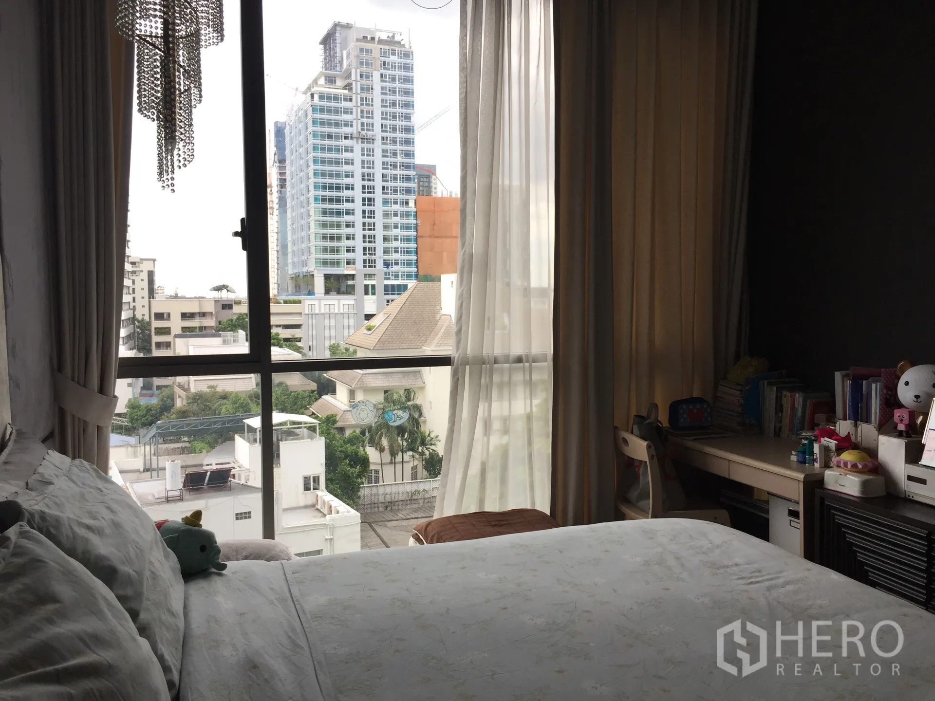 Condominium for sale in Watthana, Bangkok - Cozy bedroom with bed, study desk, sheer curtains, and floor-to-ceiling window overlooking the city.
